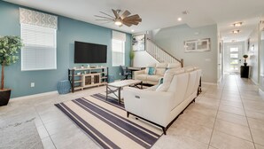 A spacious living room with a white sofa, a coffee table, and a TV. There is a staircase leading to another level, and a hallway with a door at the end. - 14 Mi to Wdw: Family-friendly Home w/ Movie Room! (Kissimmee)