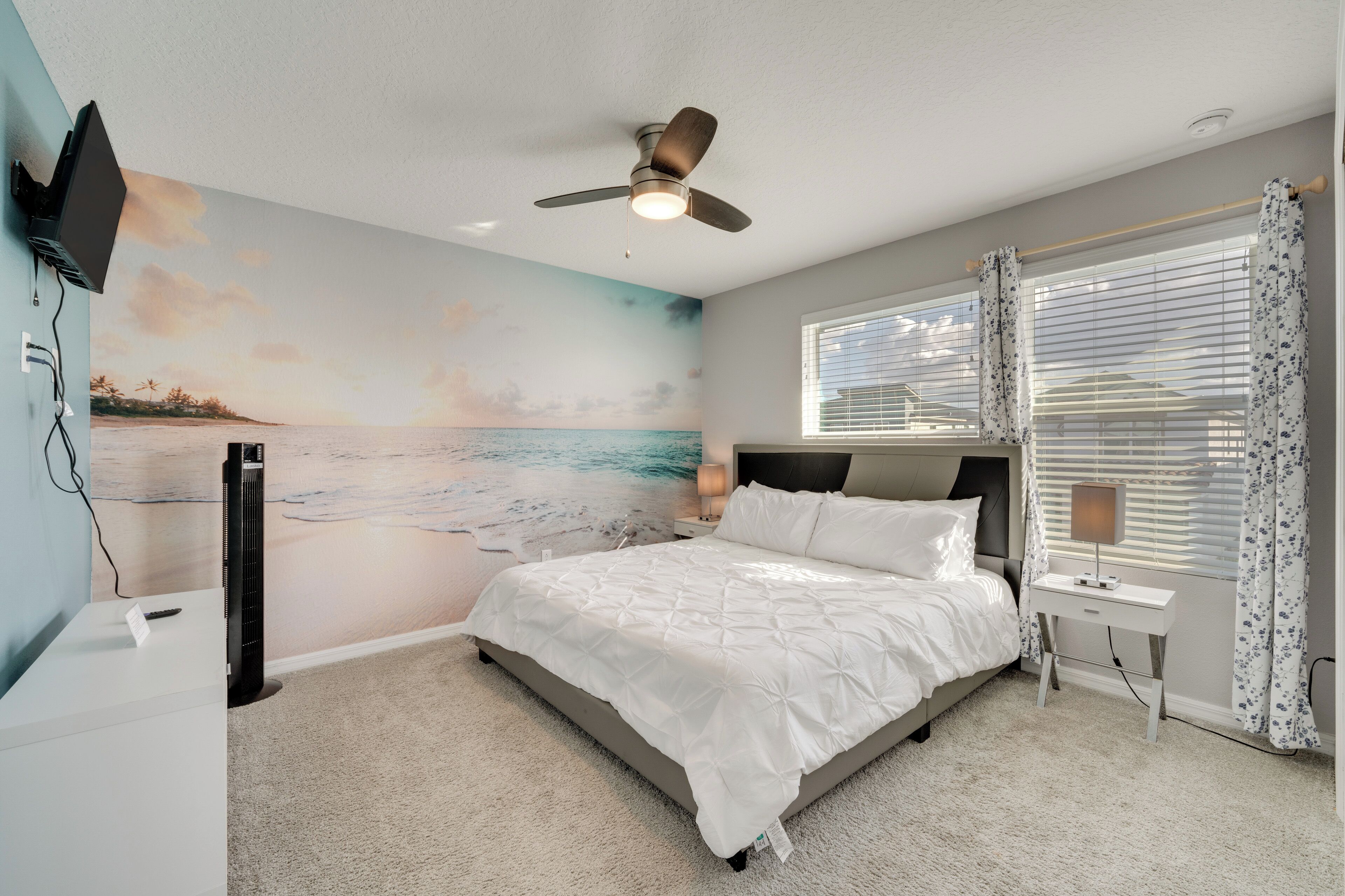 Une chambre avec un lit, un ventilateur de plafond, un téléviseur fixé au mur, une fenêtre avec des rideaux et une fresque murale représentant une plage.