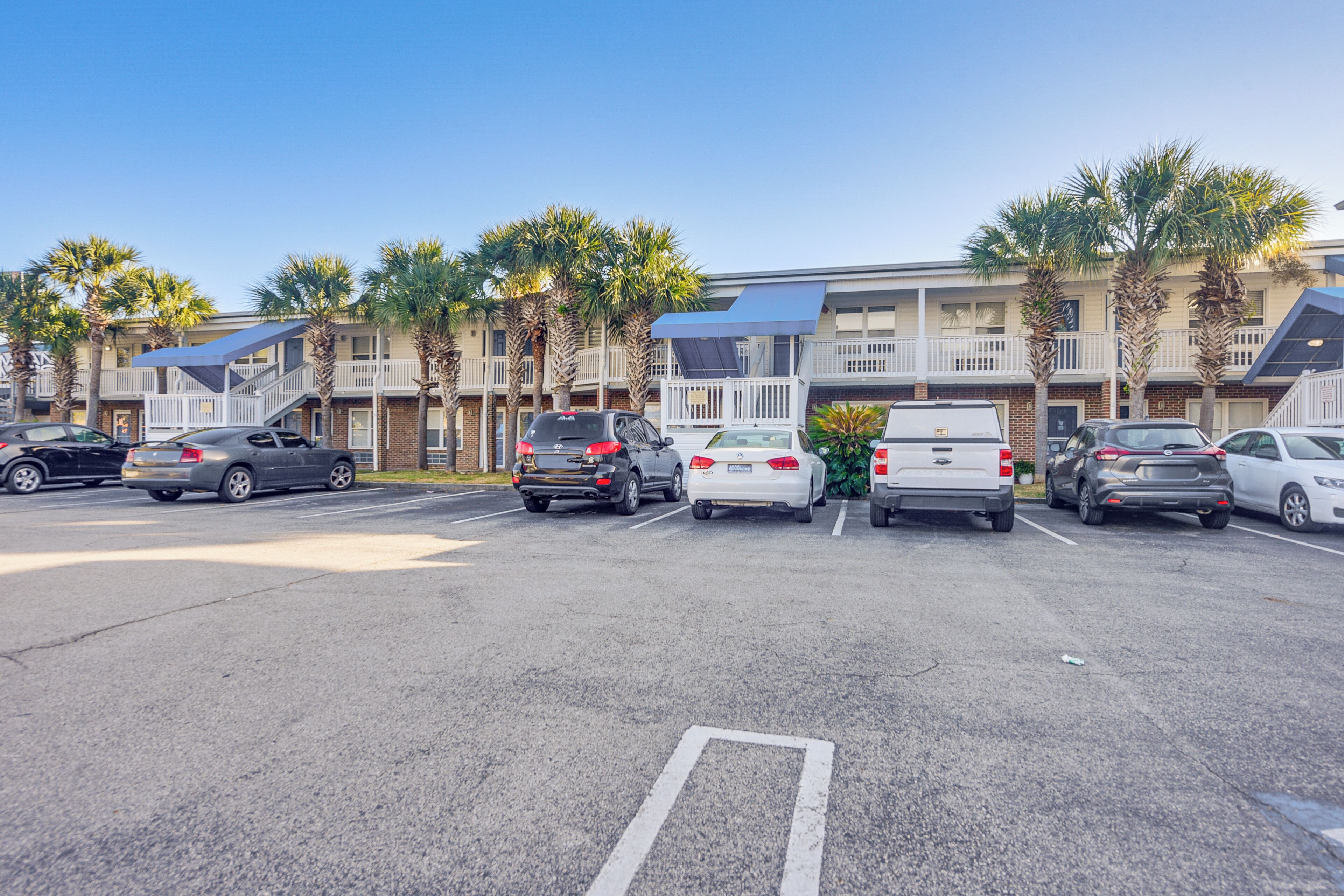 A parking lot with several cars, a building with balconies and palm trees, and a clear blue sky.