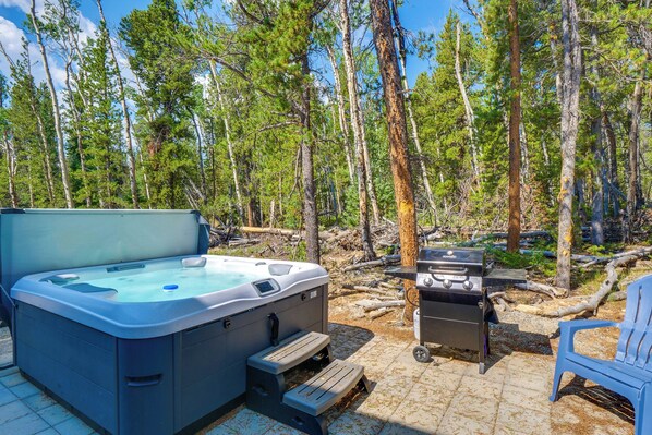 Un jacuzzi y una parrilla en un área boscosa, con una terraza de madera y una silla azul.