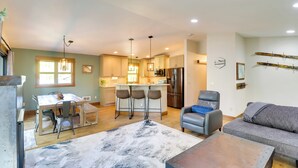 A modern kitchen with a central island, bar stools, and a dining table. There is a gray sofa and a coffee table in the living area.