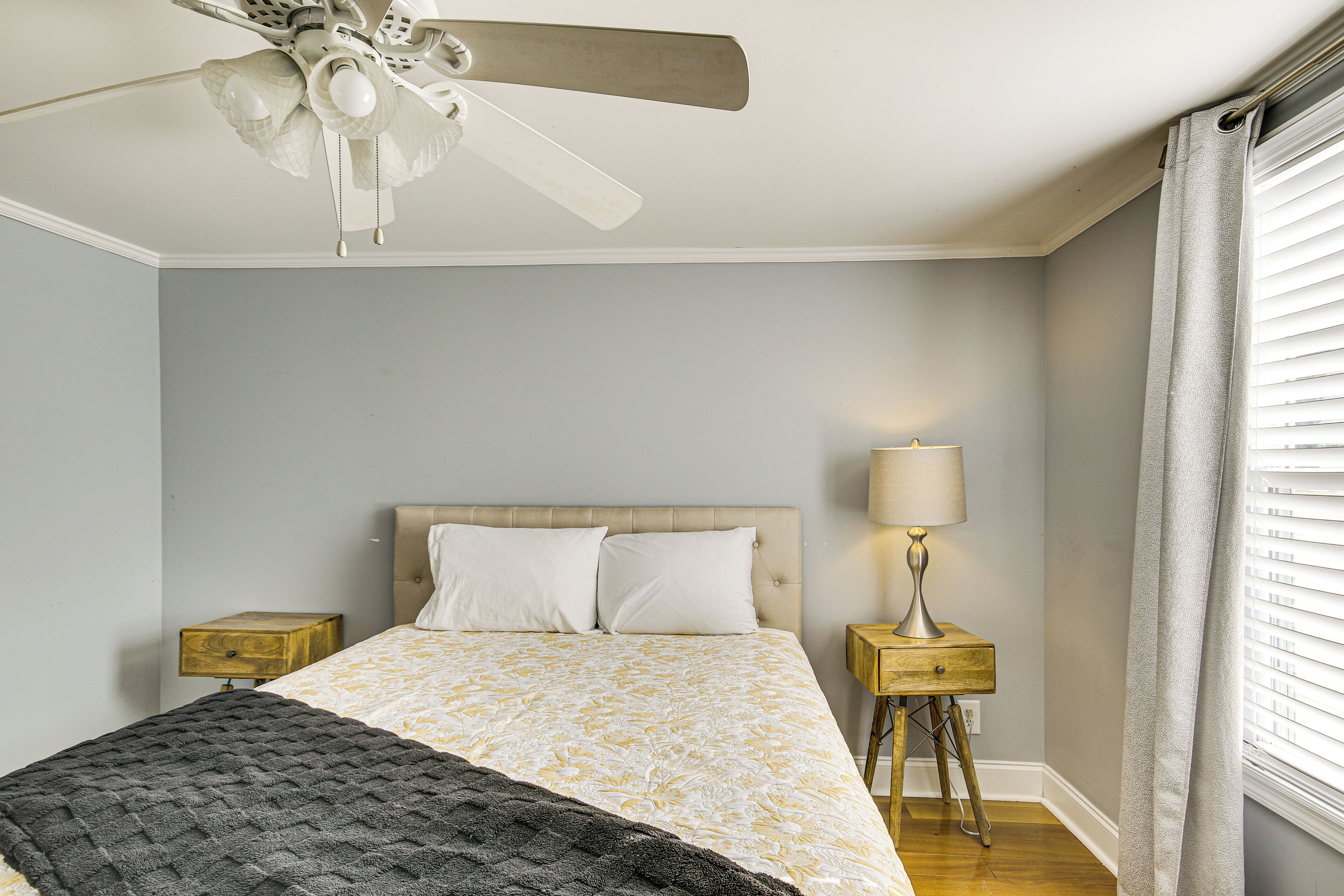 A bedroom with a bed, a ceiling fan, a bedside table with a lamp, and a window with curtains.