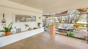 Un patio coperto con pavimento in cemento, una parete bianca con una grande finestra e un’area salotto con divano e poltrone bianchi.
