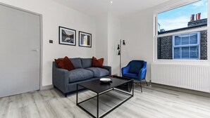 Un salon moderne avec un canapé gris, un fauteuil bleu, une table basse noire et une grande fenêtre donnant sur des immeubles.