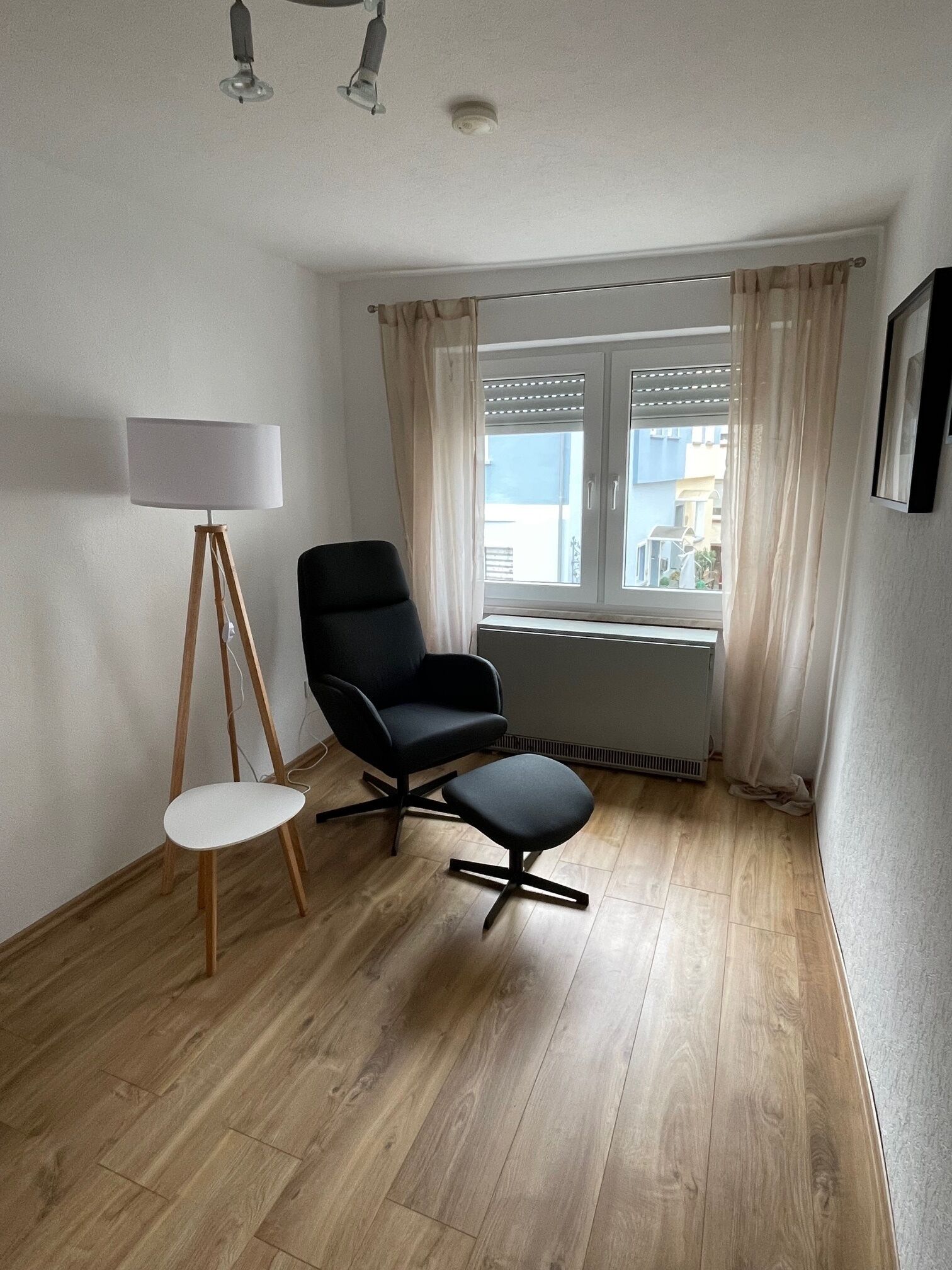 A room with a wooden floor, a black chair, a small white chair, a floor lamp, and a window with curtains.