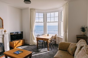A living room with a bay window overlooking the sea, a wooden coffee table, a sofa, and a television. - Beach side, newly renovated apartment with fantastic sea views. (Weymouth)