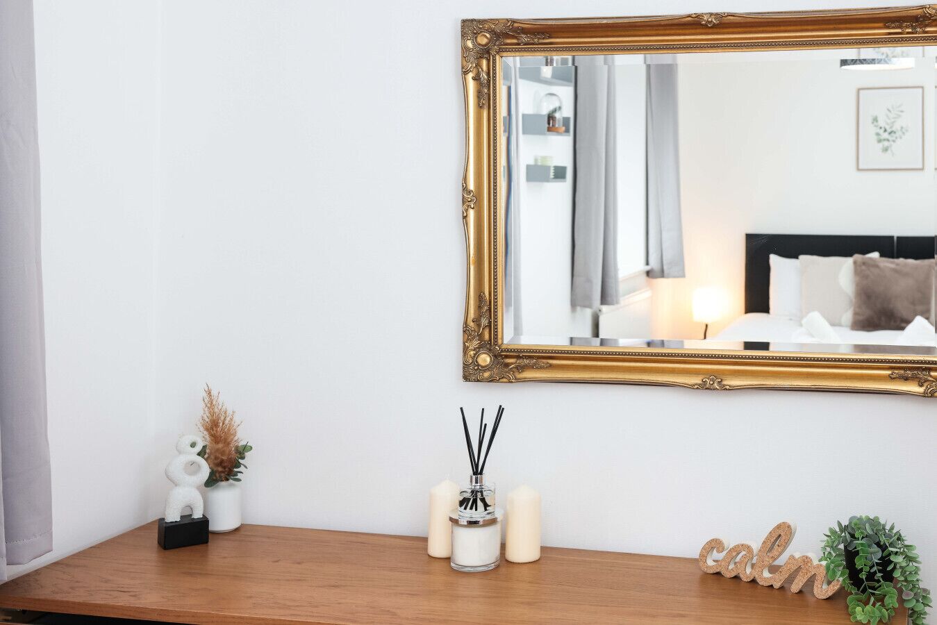 A mirror with an ornate gold frame, a wooden console table with candles, a small plant, and a decorative word 'calm' on the wall.