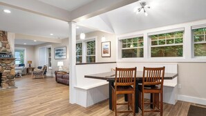 Un coin repas avec une table et des chaises en bois, une cheminée en pierre et une vue sur l’extérieur grâce à de grandes fenêtres.