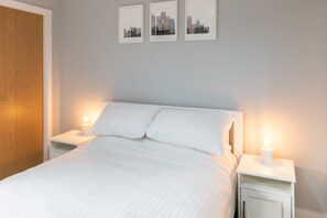 A neatly made bed with white linens, two bedside tables with lamps, and three framed cityscape pictures on the wall. - Host Stay Burton Road (Manchester)