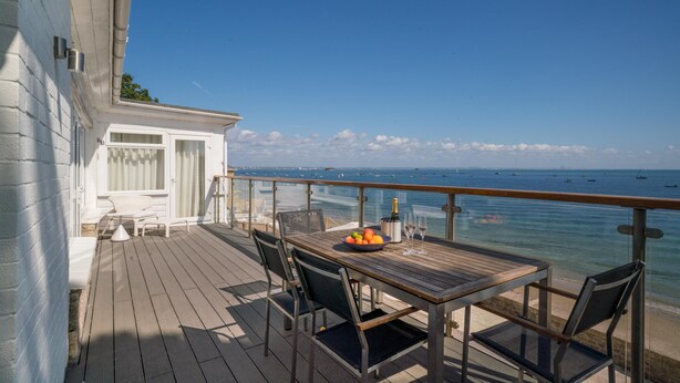 Une terrasse en bois avec une table et des chaises, d’où l’on peut admirer une mer bleue et limpide parsemée de bateaux.