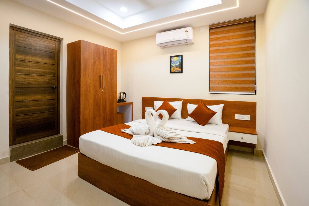 A hotel room with a wooden wardrobe, a bed with white bedding and brown pillows, a wall-mounted air conditioner, and a window with blinds.