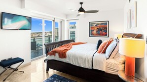 A modern bedroom with a bed, a standing lamp, a ceiling fan, and a large window offering a city view. - Charming 2-bedroom corner deluxe condo in Cupecoy with ocean and sunset view  (Cupecoy, St. Maarten)