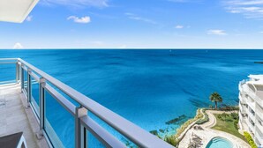 A balcony with a clear view of the ocean and a resort area with a pool and palm trees. - Charming 2-bedroom corner deluxe condo in Cupecoy with ocean and sunset view  (Cupecoy, St. Maarten)
