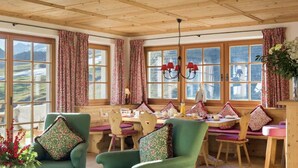 Une pièce chaleureuse avec des poutres apparentes en bois au plafond, de grandes fenêtres ornées de rideaux à motifs floraux, une table à manger dressée pour quatre personnes et des fauteuils verts recouverts de tissu, agrémentés de coussins à motifs.