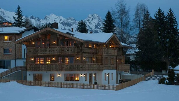 Un chalet en bois à plusieurs étages, doté de balcons, qui se dresse sur fond de montagnes enneigées.