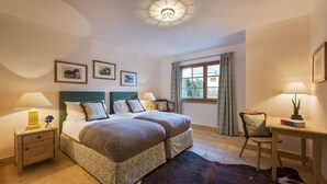 Une chambre d’hôtel avec deux lits, un bureau, une chaise, une fenêtre avec des rideaux et des tableaux encadrés au mur.