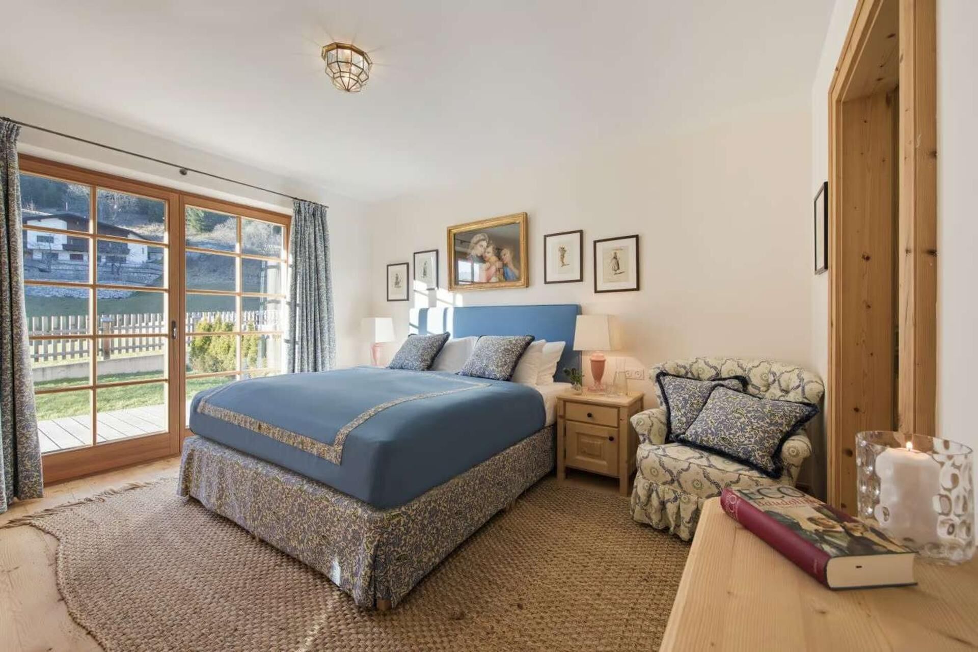 A bedroom with a large bed, a wooden nightstand, a chair, a table with a book and candle, and a view of the outdoors through a sliding door.