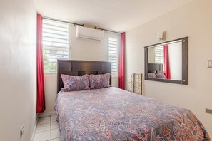 A bedroom with a bed, a window with blinds, a mirror, and an air conditioning unit.