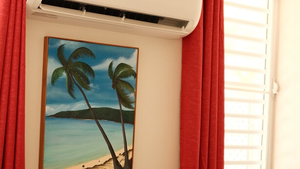 A room with a brown sofa, a painting of palm trees and a beach, red curtains, and an air conditioning unit. - Ginger House. Charming 3-bedroom townhome, fast WiFi, A/C in enchanting Rincón (Rincón)