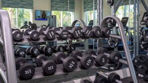 A gym with rows of dumbbells, exercise equipment, and a TV screen in the background.