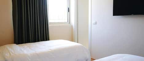 Ein Schlafzimmer mit zwei Betten, einem Fenster mit Vorhängen, einem Kleiderschrank und einem an der Wand befestigten Fernseher.
