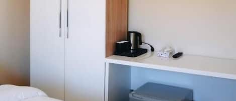 A hotel room with a bed, a wardrobe, a small desk with a coffee maker, and a trash bin.