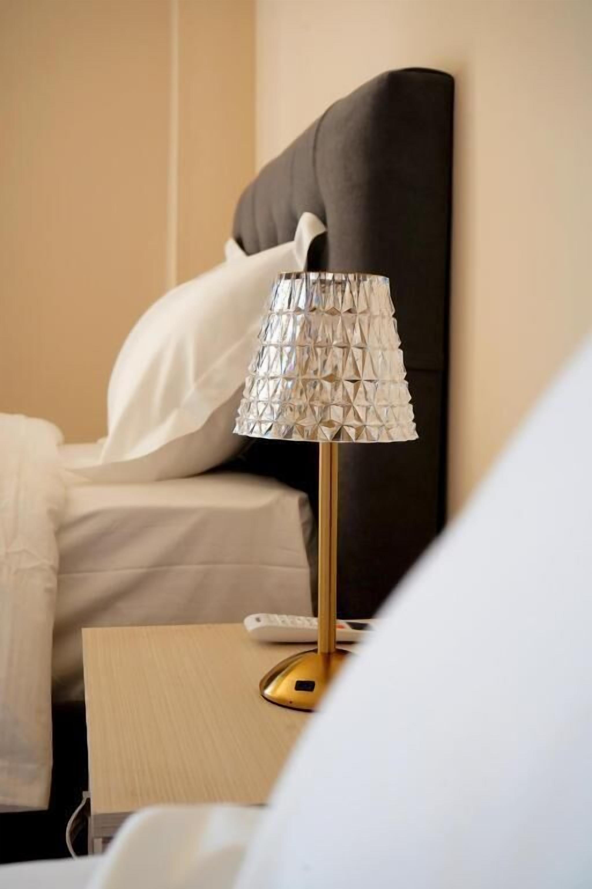 Un lit avec une couette blanche et une tête de lit noire. On peut voir une table de chevet avec une lampe et une télécommande.