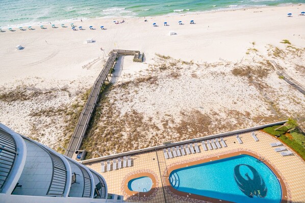 Vue aérienne d’une plage avec une promenade en bois, un espace piscine avec des chaises longues et des parasols.
