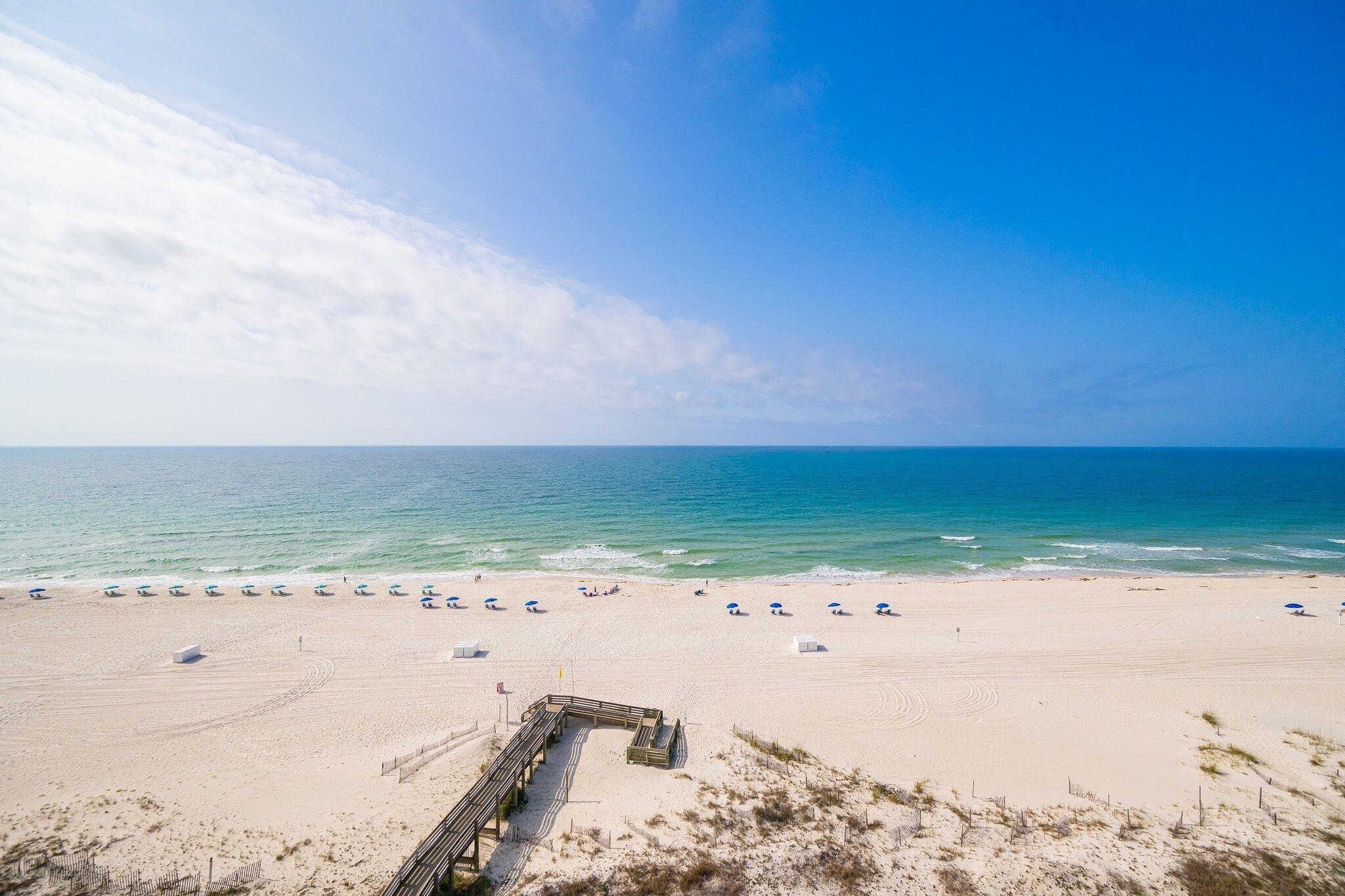 A beach with a wooden walkway leading to the water, beach umbrellas, and a clear sky.