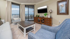 Een woonkamer met een witte bank, een gestreept fauteuil, een houten salontafel, een televisie en een kast. Er is een balkon met uitzicht op de oceaan.