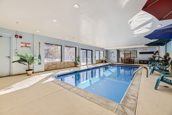 An indoor swimming pool with clear glass walls, potted plants, and sunlit windows.
