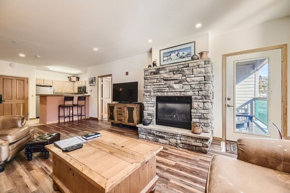 A living room with a stone fireplace, a television, and a kitchen area in the background.