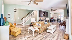 A spacious living room with a wooden floor, a staircase, and a kitchen in the background. - Lilys Pad Home with Gourmet Kitchen, Near Beach (Cape Charles)