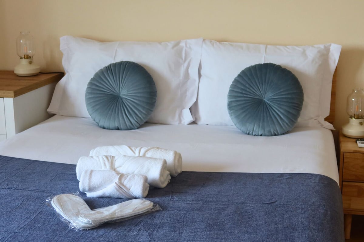 Een net opgemaakt bed met witte lakens, twee grijze kussens en een opgevouwen handdoek op een blauwe sprei.