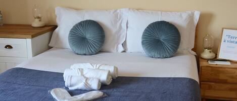 Een net opgemaakt bed met witte lakens, twee grijze kussens en een opgevouwen handdoek op een blauwe sprei.