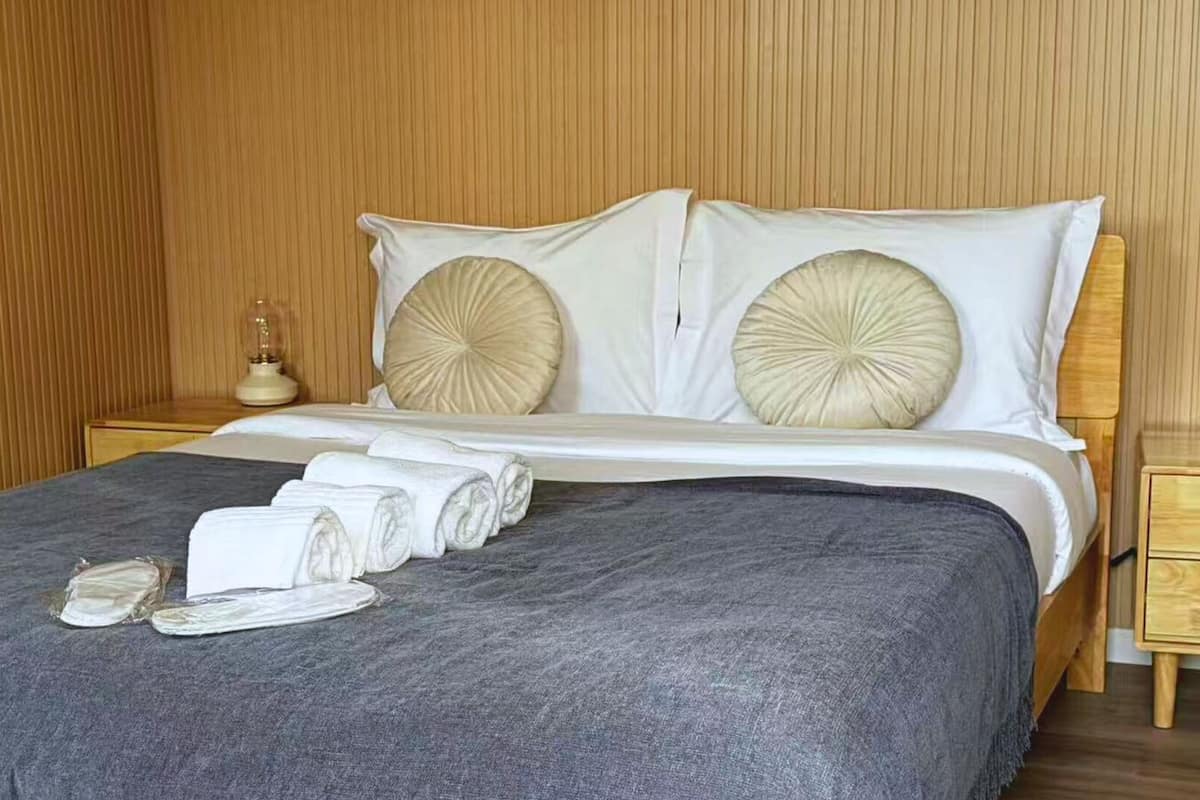 Een net opgemaakt bed met een grijze plaid, twee beige kussens en opgerolde witte handdoeken op het bed.