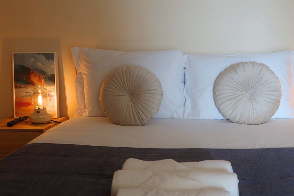 Een net opgemaakt bed met twee grote decoratieve kussens, een afstandsbediening, een ingelijste foto en een bedlampje op een houten nachtkastje.
