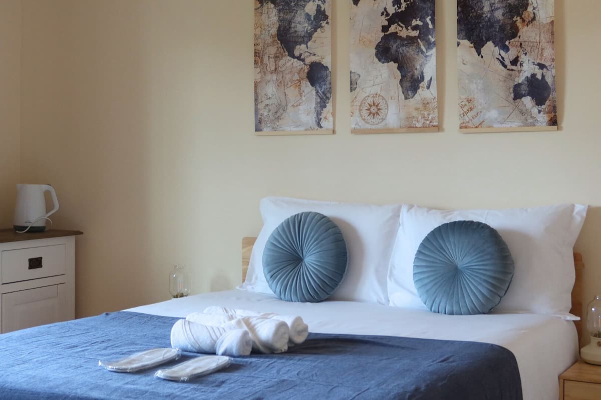 Een net opgemaakt bed met blauwe kussens, een blauwe sprei en een witte dekbedovertrek. Er staan twee nachtkastjes met lampen en een kleine vaas. Een houten dressoir met waterkoker en kopjes is zichtbaar.