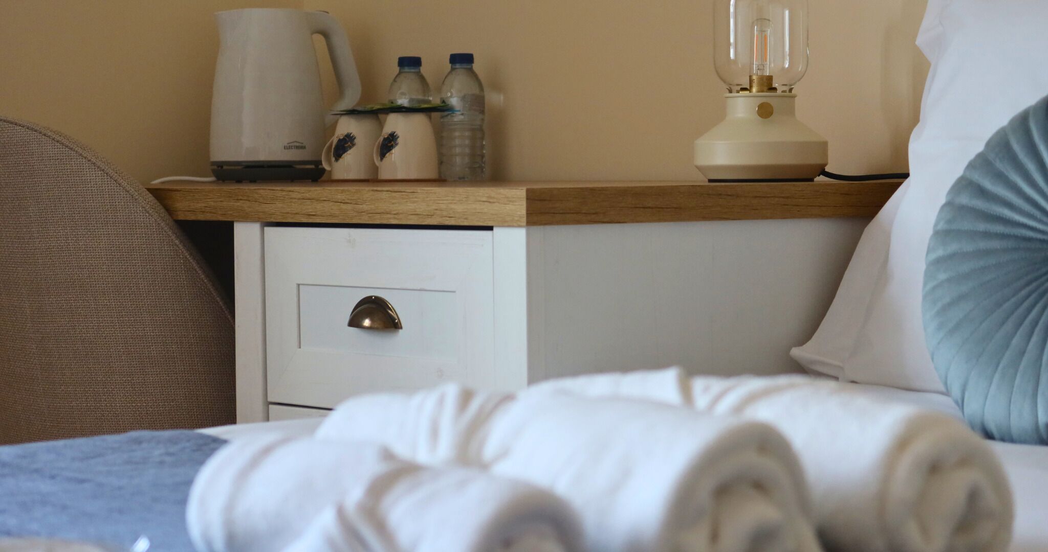A neatly made bed with white linens, a wooden nightstand with a lamp, a water bottle, and a coffee maker.