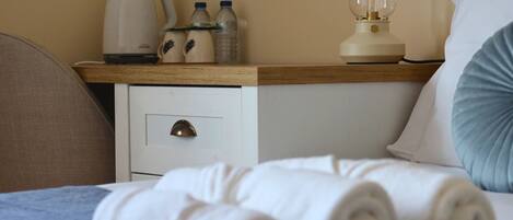 A neatly made bed with white linens, a wooden nightstand with a lamp, a water bottle, and a coffee maker.