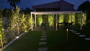 Een overdekt buitengebied met zitplaatsen, een tafel en stoelen, omgeven door een tuin met een haag en bomen.