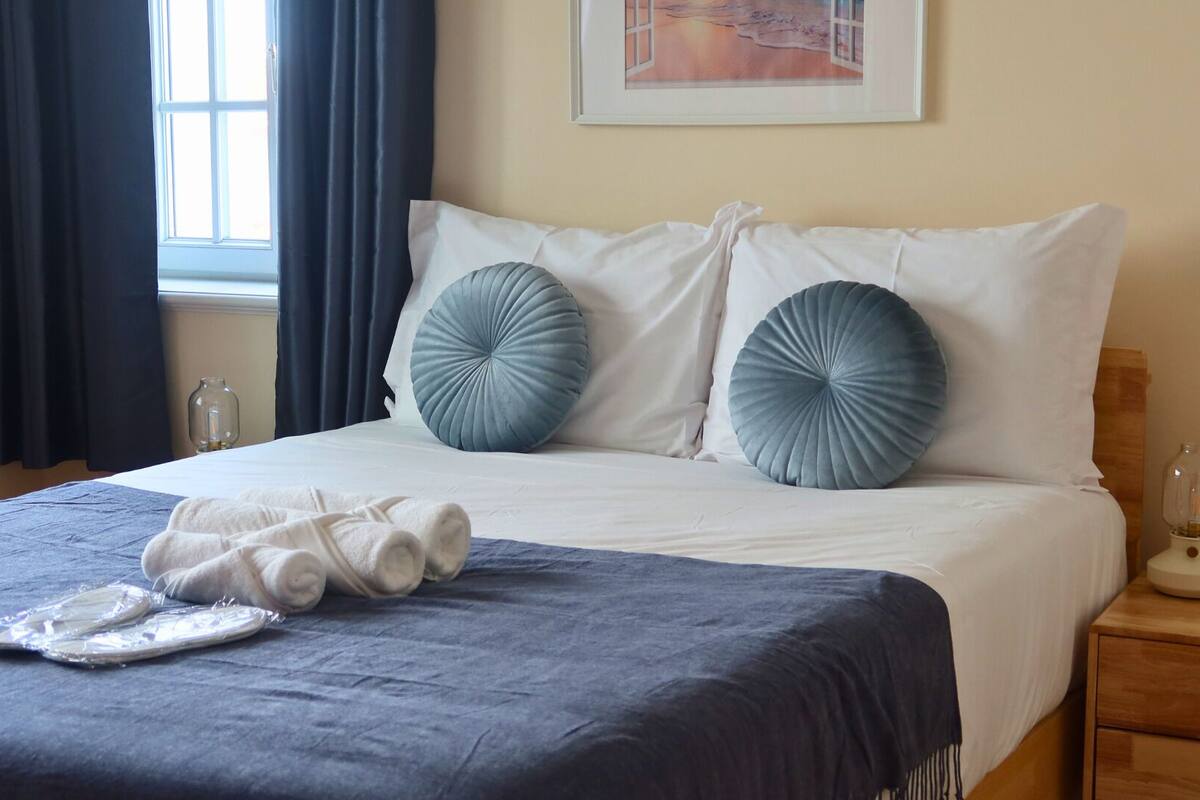 Een netjes opgemaakt bed met een blauwe sprei en twee grote, ronde, blauwe kussens. Op het bed liggen twee opgerolde handdoeken, en een klein tafeltje met een lamp en een ingelijste foto ernaast.
