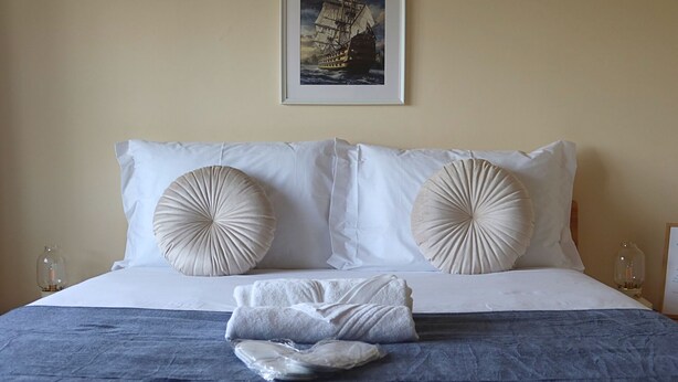 Een net opgemaakt bed met twee grote decoratieve kussens, een lijst met een foto erboven en een klein tafeltje met een glas erop.