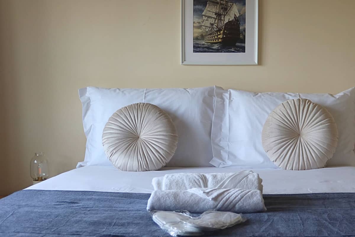 Een net opgemaakt bed met twee grote decoratieve kussens, een lijst met een foto erboven en een klein tafeltje met een glas erop.