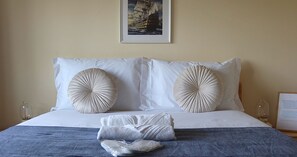 A neatly made bed with two large decorative pillows, a framed picture above the bed, and a small table with a glass on it. - Art 4 You Carcavelos Suites (Cascais)
