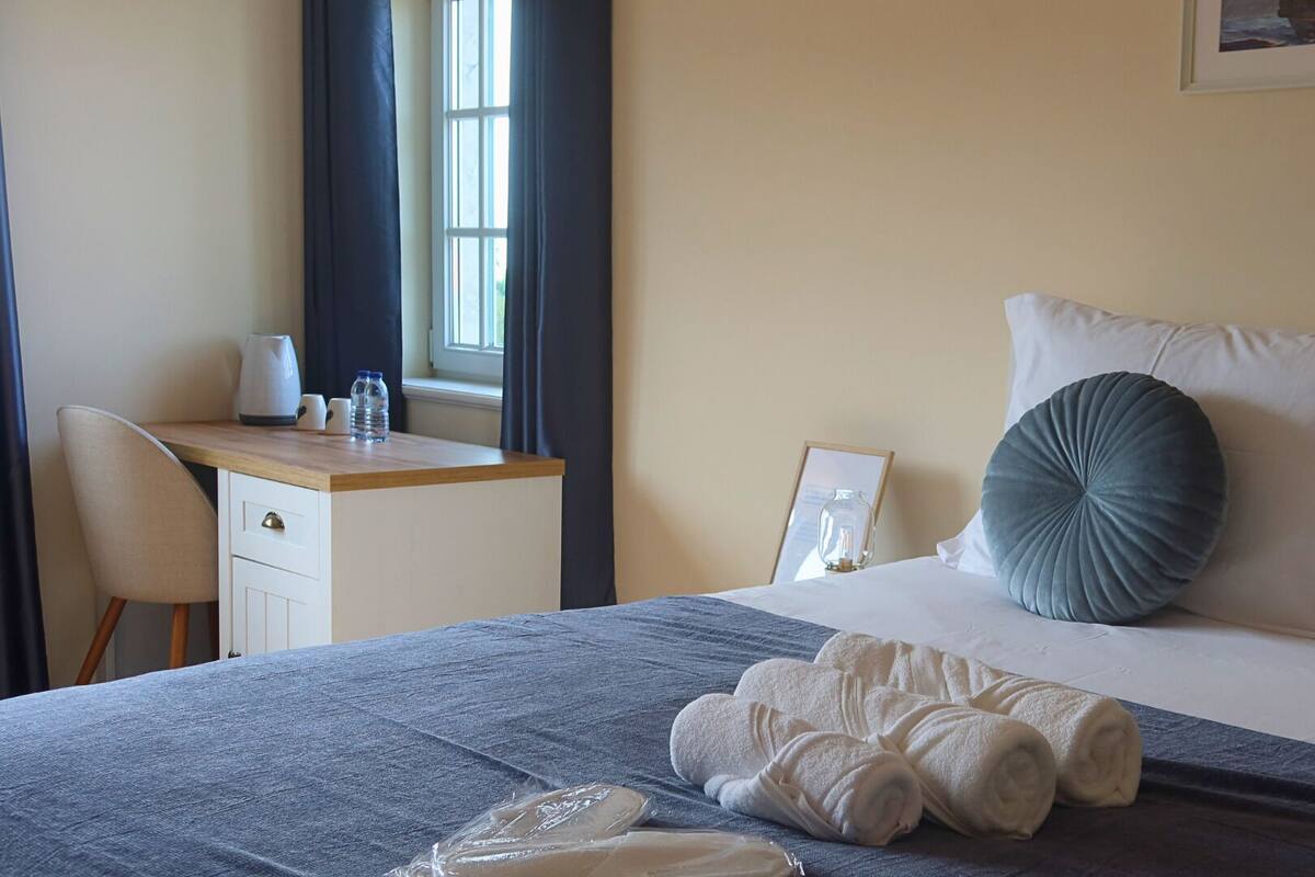 Een netjes opgemaakt bed met een grijze deken, twee kussens en opgerolde handdoeken. Naast een raam met gordijnen staat een houten bureau met een waterkoker en waterflessen.