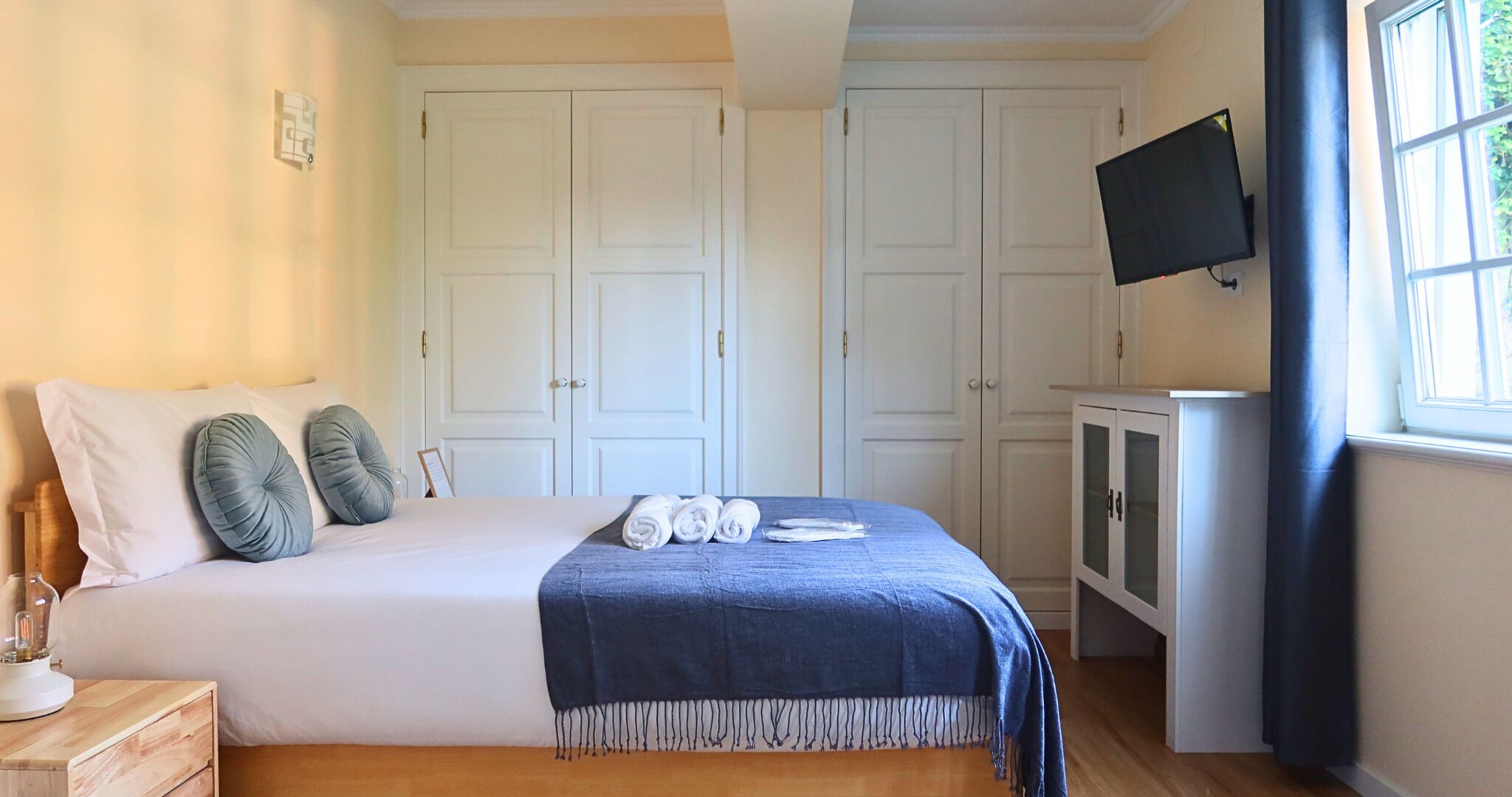 A bedroom with a bed, two pillows, a blue blanket, a wooden nightstand, a glass of water, a television on the wall, and a window with curtains.