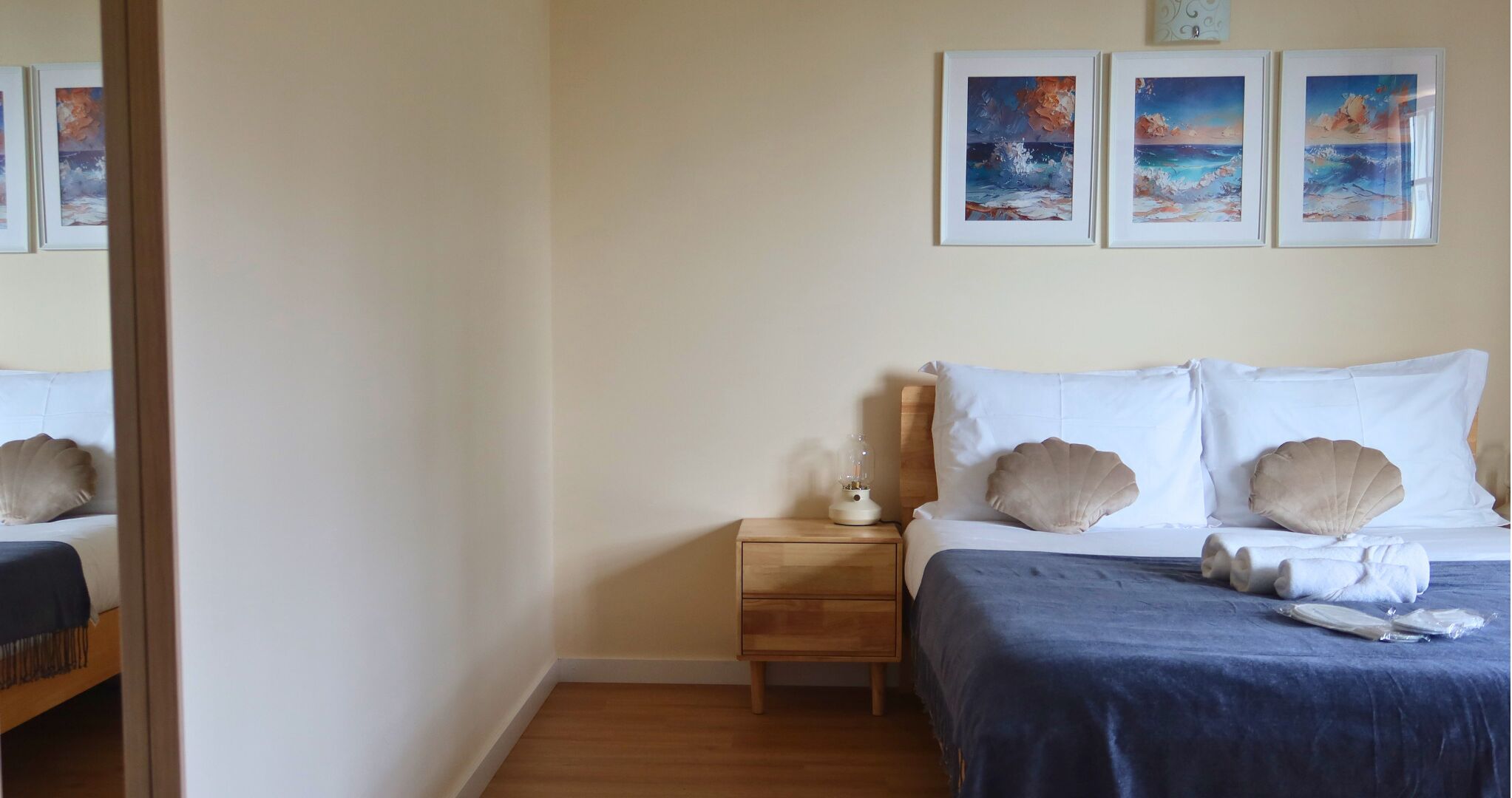 Un dormitorio con una mesita de noche de madera, una lámpara y una cama con ropa de cama blanca y almohadas con forma de concha marina. Hay tres cuadros enmarcados en la pared sobre la cama.