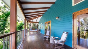 A covered porch with wooden flooring, a blue exterior wall, and a view of greenery. - Casa Mariposa (Riordanvale)