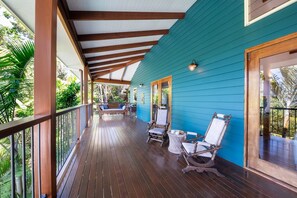 A covered porch with wooden flooring, a blue exterior wall, and a view of greenery. - Casa Mariposa (Riordanvale)